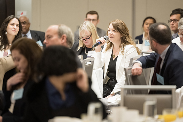 A woman surrounded by others asking a question while holding a microphone at a conference